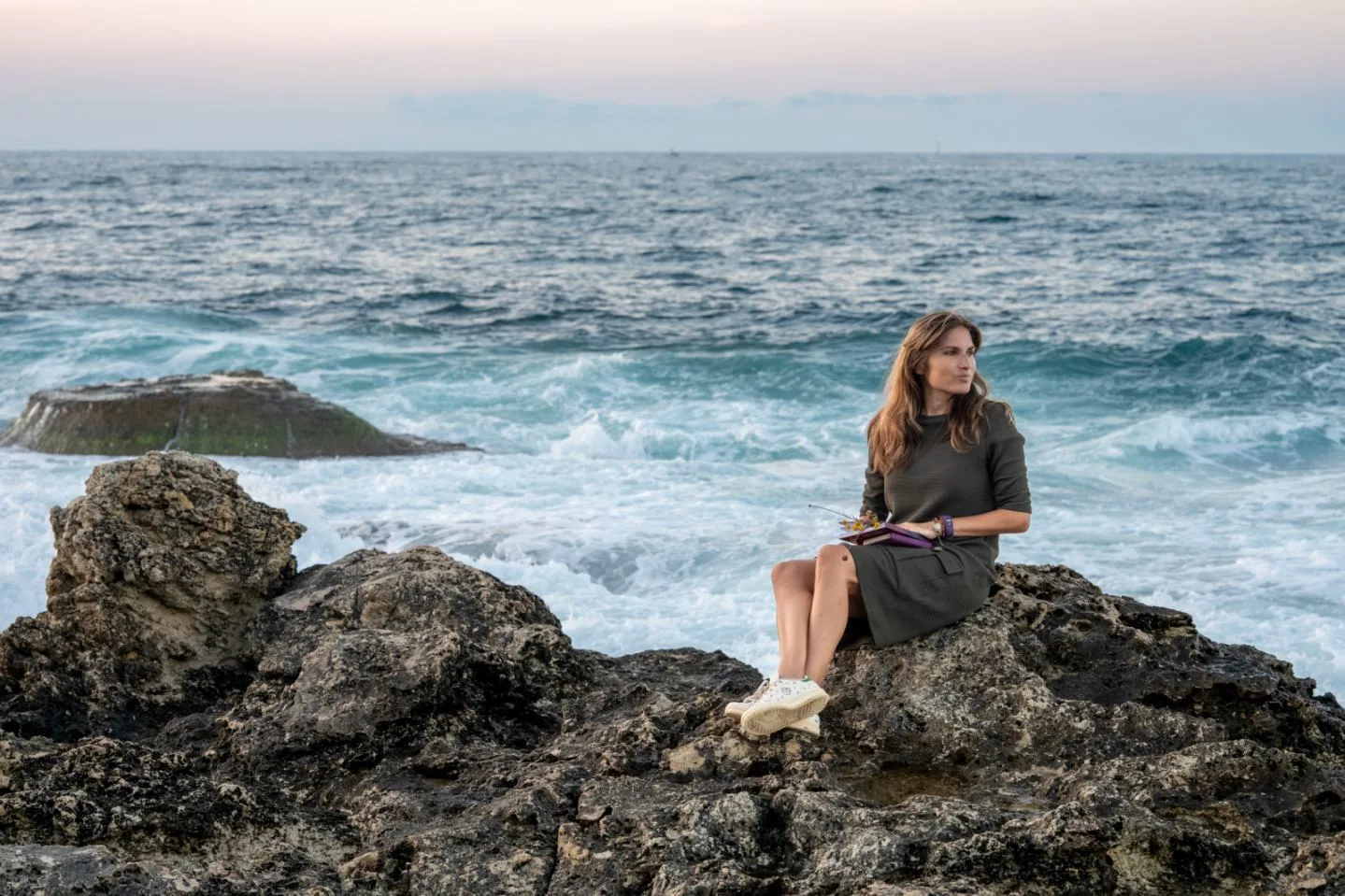 Marta holding a journal, seated on the rocky shore in front of the waves.